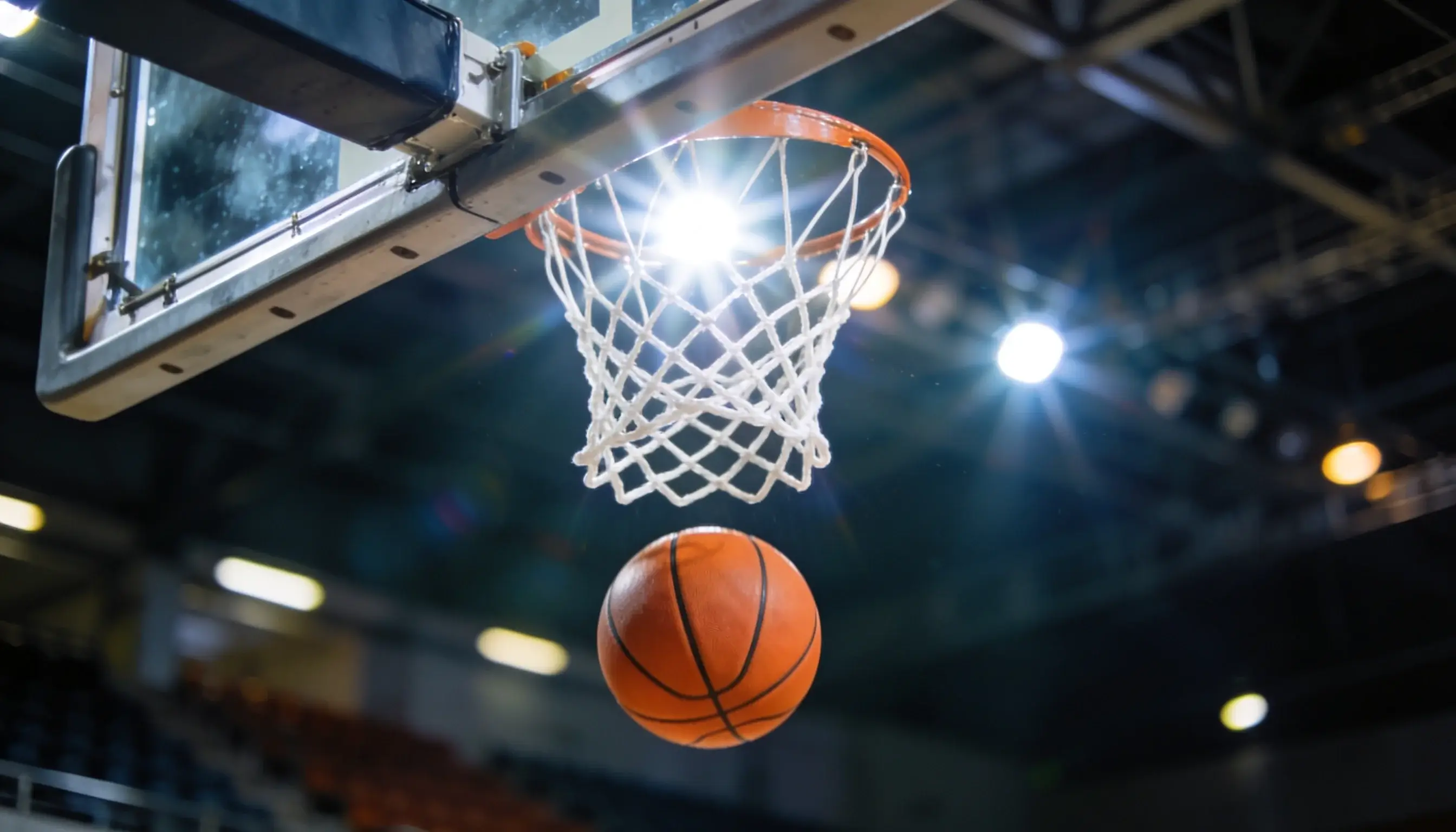 Close-up of a basketball going through a hoop net with arena lights and scoreboard in the background